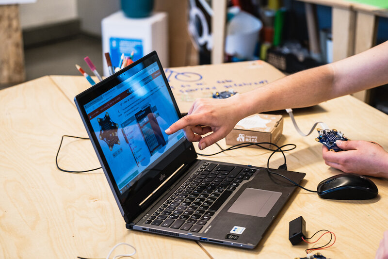 Eine Hand zeigt auf einen Laptop. Auf dem Bildschirm ist ein Calliope mini Controller und eine Programmierumgebung zu sehen.