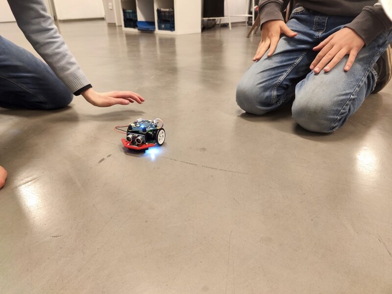 Zwei Kinder hocken auf dem Boden. Vor ihnen fährt ein selbstgebauter Roboter.