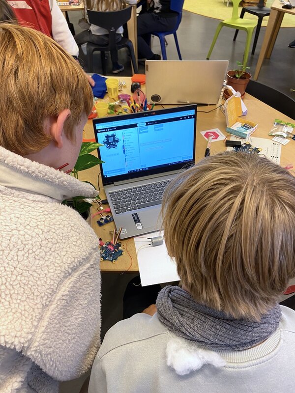 Zwei Kinder schauen sich auf einem Laptopbildschirm einen Programmcode an. Vor ihnen liegen ein Calliope Mikrocontroller und verschiedene Kabel.